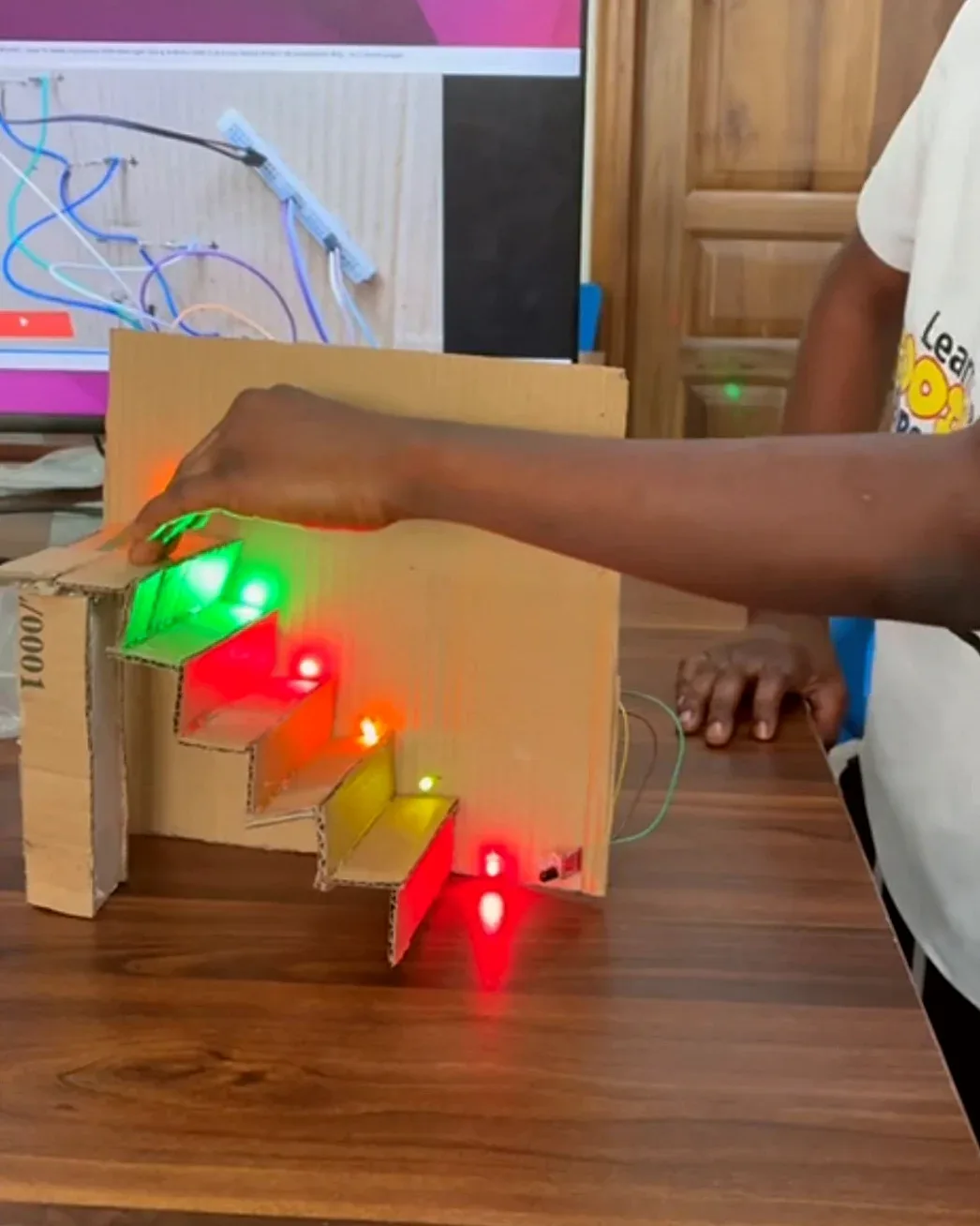 A cardboard staircase model with red and green LEDs on the steps, and a student's hand touching it